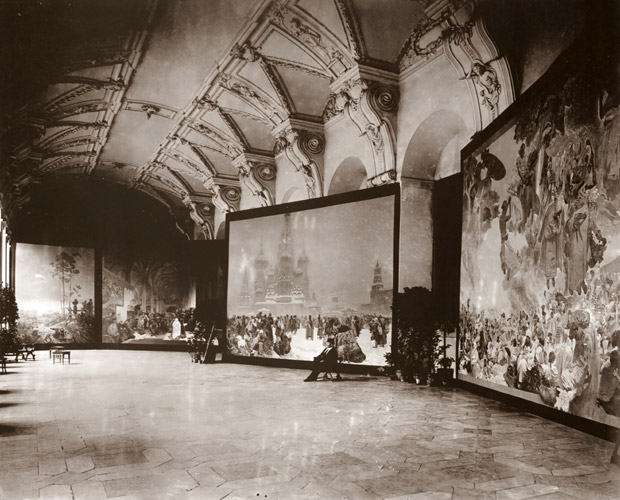 「スラヴ叙事詩」 11点の初の展覧会の記念写真。中央に座っているのがミュシャ。背後は、第6作「ロシアにおける農奴制廃止」 。 [アルフォンス・ミュシャ, 1919年, 「ミュシャが愛した光と時代」展より] パブリックドメイン画像 