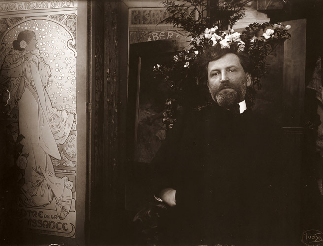アトリエの椅子に座るミュシャ。左は「椿姫」のボスター [アルフォンス・ミュシャ, 1899年頃, 「ミュシャが愛した光と時代」展より] パブリックドメイン画像 