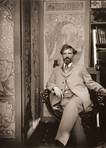 「ジスモンダ」（1894年）のボスターの前に座っているミュシャ。左には「椿姫｣（1896年）のポスターがある。 [アルフォンス・ミュシャ, 1898年頃, 「ミュシャが愛した光と時代」展より] パブリックドメイン画像 